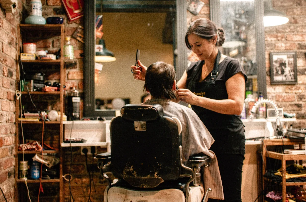 Friseurin schneidet einem Kunden im Stuhl die Haare, Kamm in der einen und Schere in der anderen Hand, im Backsteinsalon mit vollen Regalen.