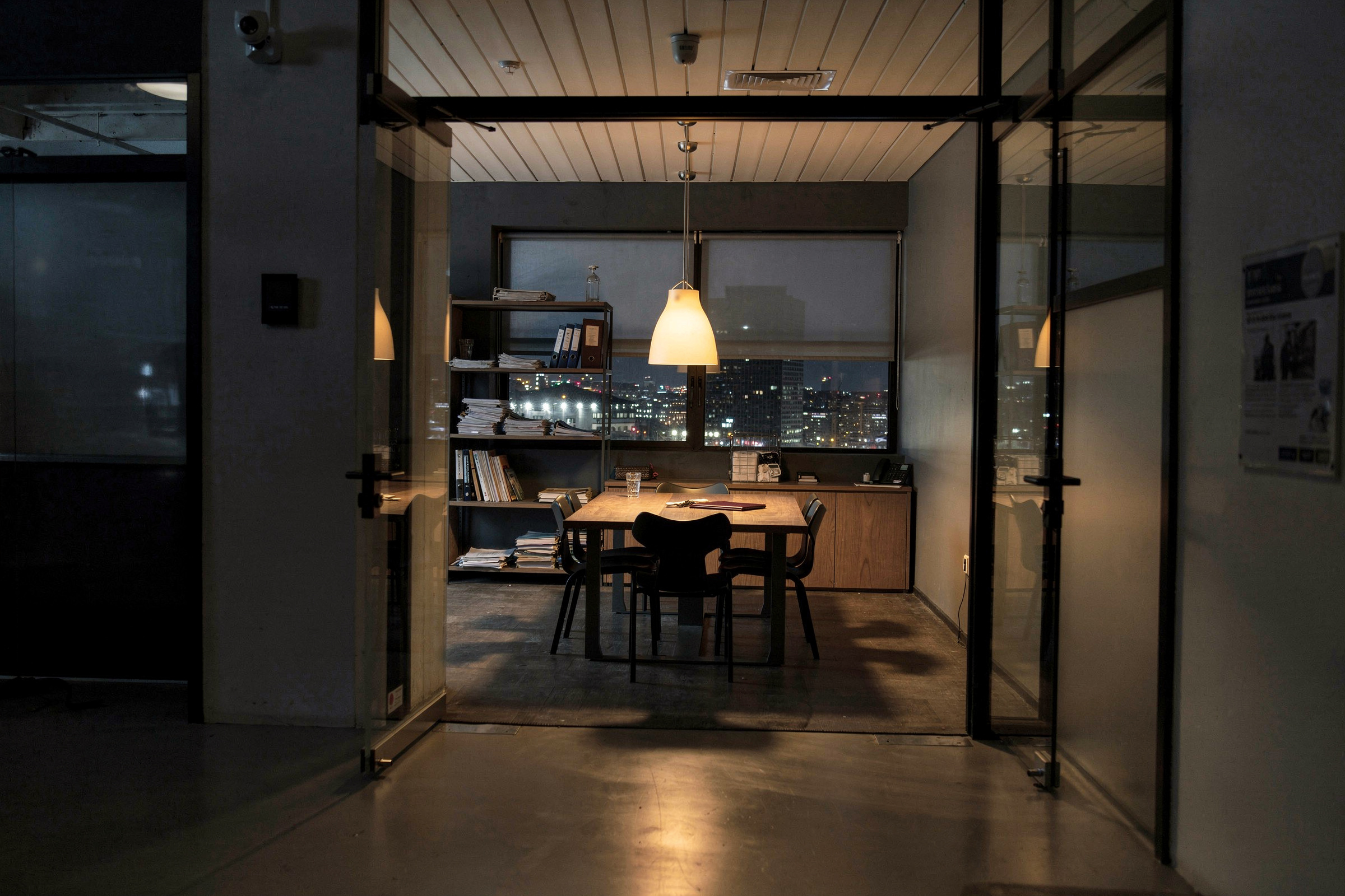 Ein leerer Besprechungsraum einer kleinen Kanzlei am späten Abend, vom Flur aus durch die Glaswand gesehen: eingeschobene Stühle, eine geschlossene Mappe auf dem Tisch und eine warme Lampe, die noch brennt.