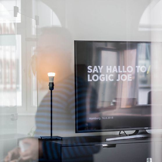 Büro-Detail durch eine Glasscheibe: Bildschirm und Stehlampe, in der Spiegelung ein Mitarbeiter