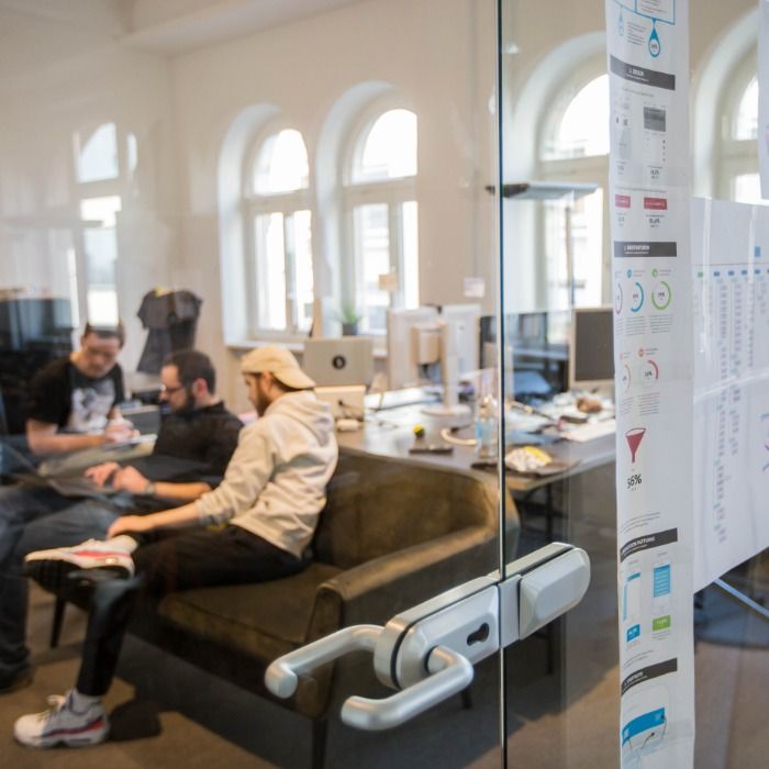 Drei Kollegen arbeiten mit Laptop auf dem Sofa, an der Glastür hängen ausgedruckte Entwürfe