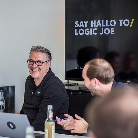 Zwei Kollegen lachen im Meeting, dahinter ein Bildschirm mit dem Logic-Joe-Slogan